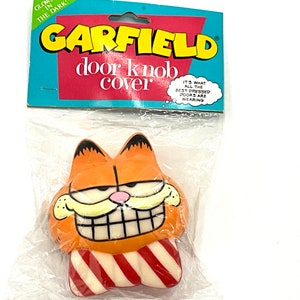 May include: A Garfield door knob cover featuring a cartoon image of Garfield wearing a red and white striped scarf. The packaging says "Glow in the dark" and "It's what all the best dressed doors are wearing."