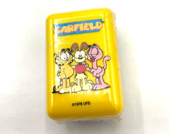 May include: A yellow plastic case with a Garfield cartoon image. The image features Garfield, Odie, and Nermal. The text "GARFIELD" is printed in black letters on the case. The copyright information "©1978 UFS" is printed in black letters on the bottom of the case.