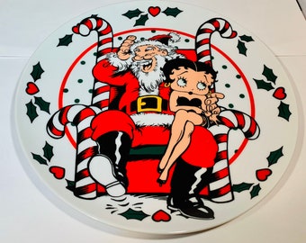 Peut inclure: Une assiette de Noël en céramique blanche avec une bordure de canne à sucre rouge et blanche. Le Père Noël est assis dans un fauteuil en canne à sucre avec Betty Boop assise sur ses genoux. L'assiette est décorée d'un motif de feuilles de houx vertes.