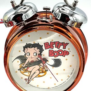 May include: A copper-colored alarm clock featuring a Betty Boop design on the clock face. The clock has a silver handle and bells, with a white clock face adorned with gold stars. The Betty Boop image is in the center, with the words "BETTY BOOP" above.