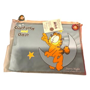 May include: A clear plastic pouch featuring Garfield and Odie. The design shows Garfield sitting on a crescent moon with stars, the text "Starry Night", and the words "Garfield and Odie". The pouch has a zipper closure and a Garfield charm.