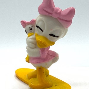 May include: A white and yellow plastic figurine of a duck with a pink bow holding a smaller duck figurine. The larger duck is standing on a yellow surfboard.