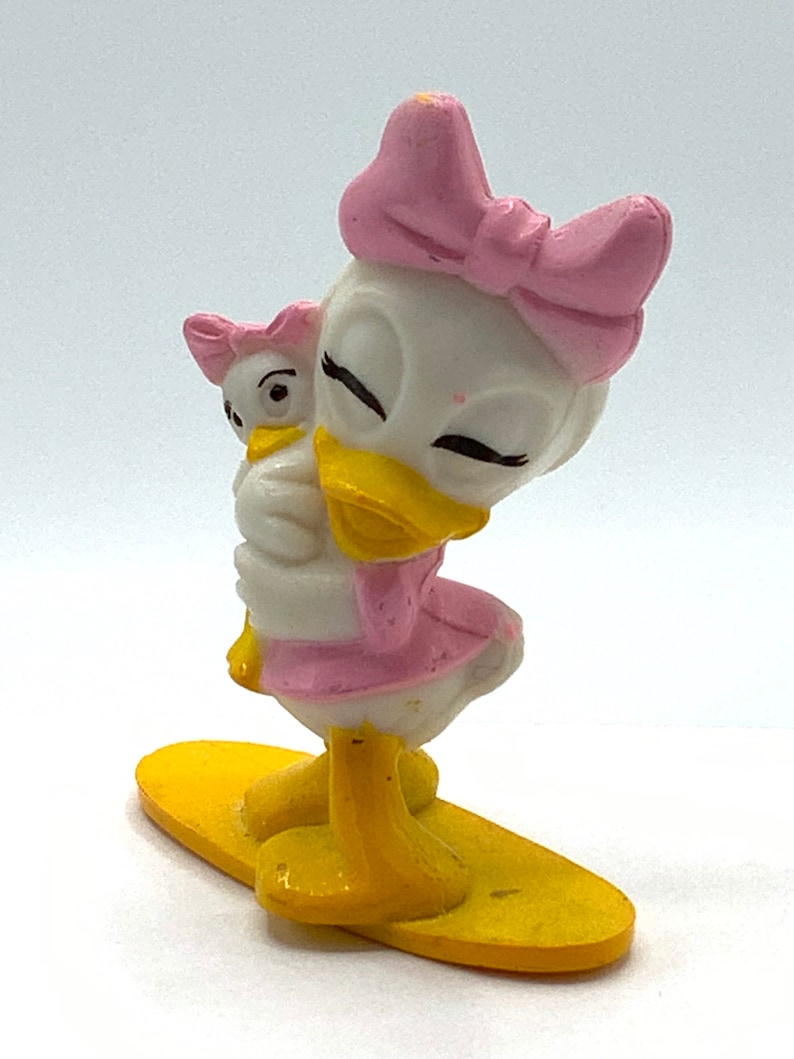 May include: A white and yellow plastic figurine of a duck with a pink bow holding a smaller duck figurine. The larger duck is standing on a yellow surfboard.
