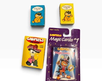 May include: A collection of Garfield card games including a blue box, a yellow box, a yellow Old Maid card game box, and a purple blister-packed set labeled Garfield Magic Cards #1, featuring illustrations of Garfield and Odie with text.