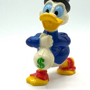 May include: A plastic figurine of a cartoon duck wearing a top hat and holding a money bag. The duck is blue, yellow, and white with a green dollar sign on the bag.