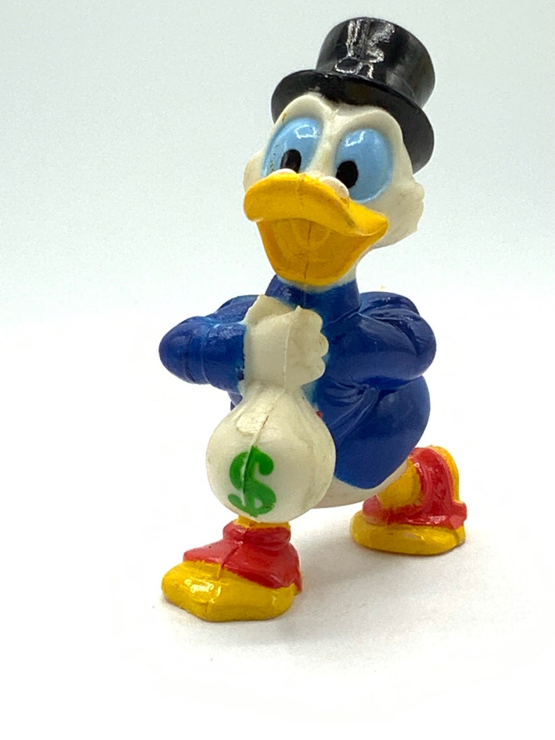 May include: A plastic figurine of a cartoon duck wearing a top hat and holding a money bag. The duck is blue, yellow, and white with a green dollar sign on the bag.