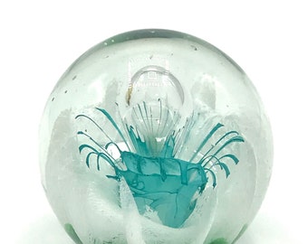 May include: A clear glass paperweight with a green and white swirl design and a teal blue flower inside.