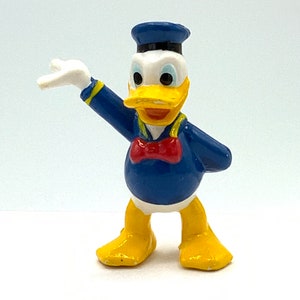 May include: A yellow and blue plastic figurine of Donald Duck wearing a blue sailor hat and a red bow tie. He is standing with his right arm raised and his left arm bent at the elbow.