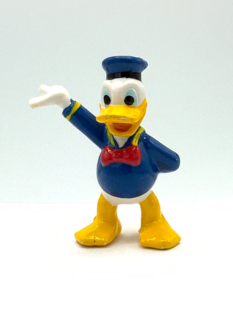 May include: A yellow and blue plastic figurine of Donald Duck wearing a blue sailor hat and a red bow tie. He is standing with his right arm raised and his left arm bent at the elbow.