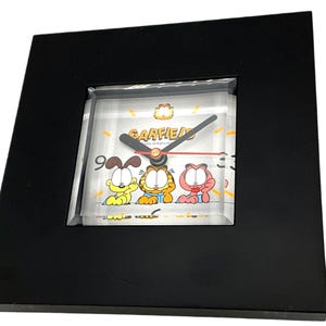 May include: Black square clock featuring the Garfield cartoon characters. The clock face displays Garfield, Odie, and Arlene, with the word "GARFIELD" above. The clock has black hands and a red second hand. The clock is encased in a black frame.