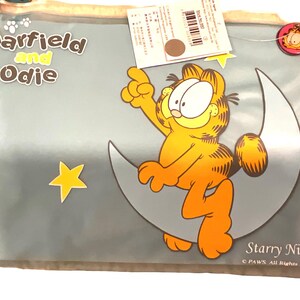 May include: A clear plastic bag featuring Garfield the cat sitting on a crescent moon with a yellow star. The words "Garfield and Odie" are at the top, and "Starry Night" is at the bottom. The cartoon cat is orange and yellow.