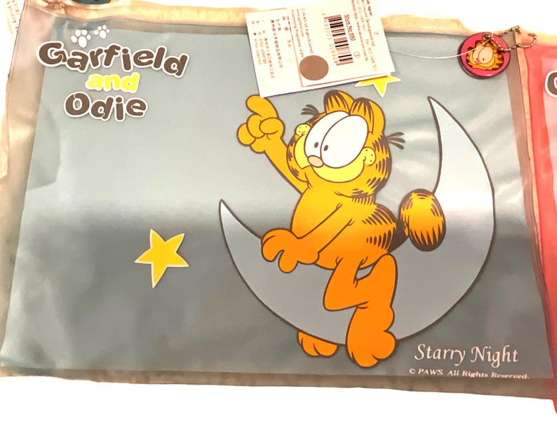 May include: A clear plastic bag featuring Garfield the cat sitting on a crescent moon with a yellow star. The words "Garfield and Odie" are at the top, and "Starry Night" is at the bottom. The cartoon cat is orange and yellow.
