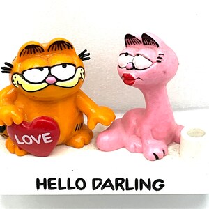 Puede incluir: Un conjunto de figuras que representa a Garfield el gato sosteniendo un corazón rojo que dice "LOVE" y una gata rosa. La base de la figura es blanca y dice "HELLO DARLING".