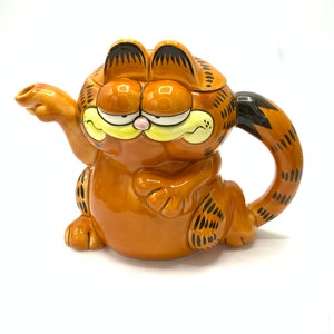 May include: A ceramic teapot shaped like Garfield the cat. The teapot is orange with black stripes and a yellow nose. The teapot has a handle and a spout.