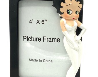 Puede incluir: Un marco de fotos rectangular negro con esquinas doradas, que presenta una figura 3D de Betty Boop con un vestido blanco y collar de perlas. El inserto blanco muestra el texto 4" x 6", Picture Frame y Made In China.