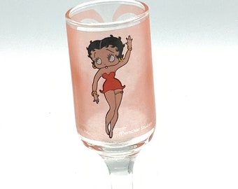 Peut inclure: Un verre à shot en verre rose avec une image de dessin animé de Betty Boop portant une robe rouge et faisant signe. Le verre a une tige et une base claires.