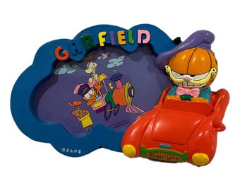 May include: A blue cloud-shaped picture frame with the word "GARFIELD" in colorful letters. The frame features a cartoon image of Garfield and friends on a train. A red car with a Garfield figure is attached to the frame.