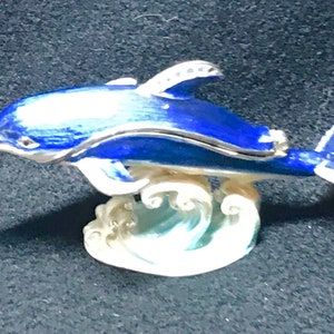 May include: A blue and silver dolphin figurine with a hinged lid, sitting on a white wave base. The dolphin is a popular symbol of good luck and is often used as a decorative item.