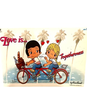 May include: A vintage 1986 Love is... print featuring a cartoon couple riding a red tandem bicycle. The text reads Love is... Togetherness by Kim Casali. The scene includes palm trees, a basket with bread and a bottle, and three red hearts.
