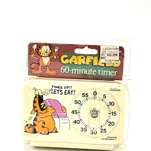 May include: A white kitchen timer featuring Garfield, a cartoon cat, with the text "Times Up!! Let's Eat!" and a 60-minute timer dial. The timer is packaged in a clear plastic bag with a price tag.