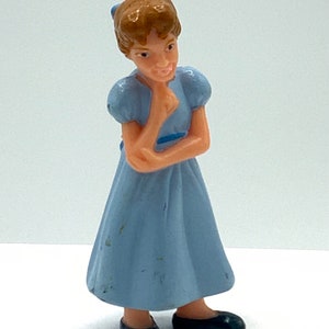 May include: A small plastic figurine of a girl with brown hair wearing a light blue dress and black shoes. She is holding her chin with her hand and looking to the side.