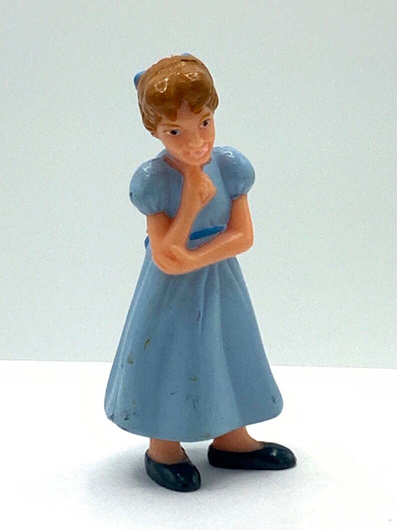 May include: A small plastic figurine of a girl with brown hair wearing a light blue dress and black shoes. She is holding her chin with her hand and looking to the side.