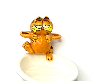 May include: A ceramic toothbrush holder featuring a cartoon orange cat with black stripes, holding a toothbrush in each hand. The cat is sitting on a white ceramic bowl.