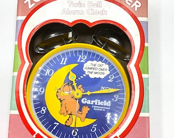 May include: A yellow Garfield twin bell alarm clock with a blue face and silver bells. The clock face features a cartoon image of Garfield sleeping on a crescent moon. The text "The cat jumped over the moon" is printed on the clock face. The text "Garfield" and "© 1978 United Feature Syndicate, Inc." is printed below the clock face.