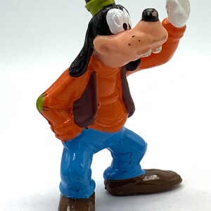 May include: A plastic figurine of Goofy, a cartoon dog character from Disney. He is wearing a green hat, an orange shirt, a brown vest, blue jeans, and brown shoes. He is standing with his right arm raised and his left hand on his hip.
