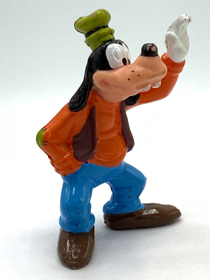May include: A plastic figurine of Goofy, a cartoon dog character from Disney. He is wearing a green hat, an orange shirt, a brown vest, blue jeans, and brown shoes. He is standing with his right arm raised and his left hand on his hip.