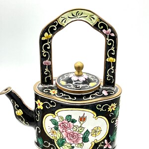 May include: A black cloisonné teapot with a floral design. The teapot has a gold handle and a gold lid. The floral design includes pink roses, yellow flowers, and green leaves.