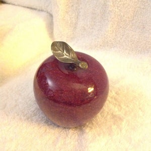 May include: A dark purple glass apple with a silver metal leaf on top. The apple is sitting on a white fluffy surface.
