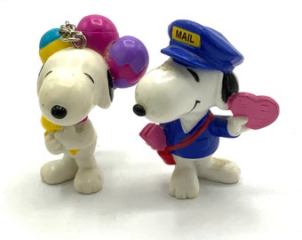 Può includere: Due piccole figurine di Snoopy in plastica. Uno Snoopy tiene palloncini blu, gialli, rosa e viola. L'altro Snoopy indossa un cappello blu con la scritta "MAIL" e tiene un cuore rosa.