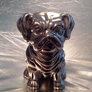 May include: A silver ceramic pug figurine with a wrinkled face and a short tail. The figurine is sitting on a white surface.