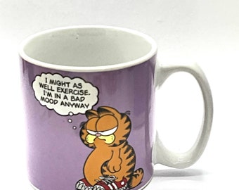 May include: A purple ceramic mug with a cartoon image of Garfield the cat. The mug has a white handle and the text "I might as well exercise. I'm in a bad mood anyway." in a thought bubble above Garfield's head.