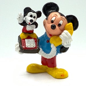 May include: A vintage figurine of Mickey Mouse on the phone. He is holding a yellow phone receiver to his ear and a smaller Mickey Mouse figurine is holding a red phone with a black keypad. The larger Mickey Mouse is wearing a blue shirt, red pants, and yellow shoes.