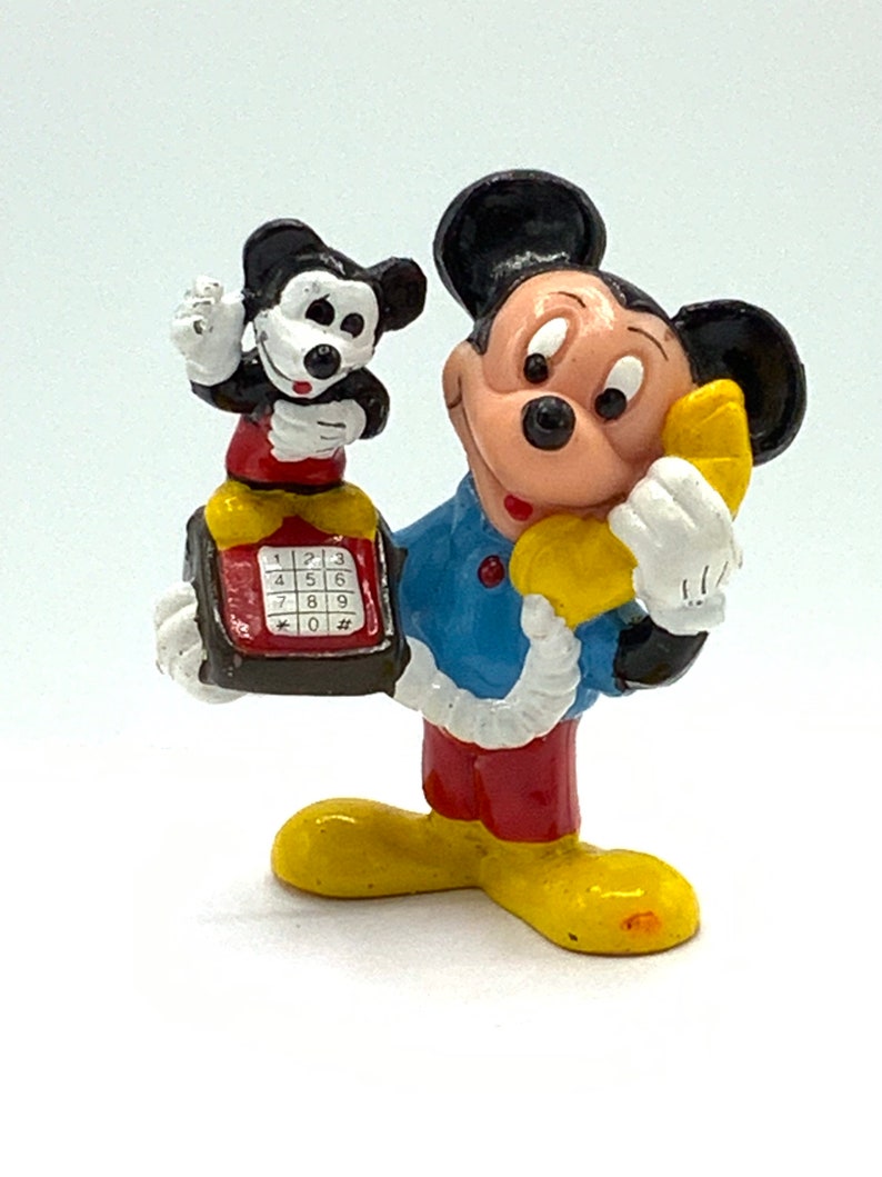 May include: A vintage figurine of Mickey Mouse on the phone. He is holding a yellow phone receiver to his ear and a smaller Mickey Mouse figurine is holding a red phone with a black keypad. The larger Mickey Mouse is wearing a blue shirt, red pants, and yellow shoes.