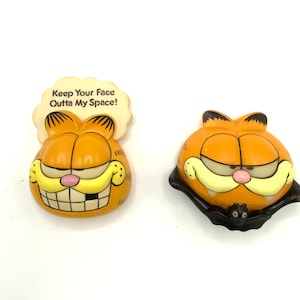 May include: Two ceramic Garfield the cat figurines. One is a standard Garfield face with the text "Keep Your Face Outta My Space!" on a speech bubble. The other is a Halloween-themed Garfield with a black bat cape and a small bat on the cape.