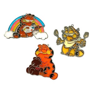 May include: Set of three Garfield enamel pins. One pin features Garfield painting a rainbow, another shows Garfield with a spoon and fork, and the last depicts Garfield hugging a teddy bear. Each pin has a silver outline.