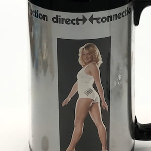 May include: A black coffee mug with a silver metallic finish. The mug features a woman in a white tank top and shorts. The text "direction direct connection" is printed on the mug.