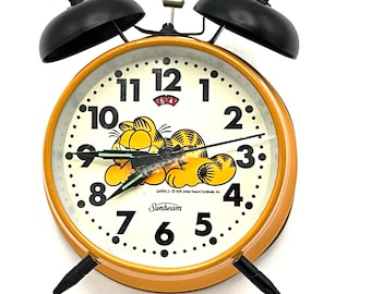 May include: A yellow and black alarm clock with a Garfield cartoon character on the face. The clock has a black metal frame and a white face with black numbers. The clock is set to 7:54. The clock is made by Sunbeam.