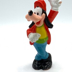 May include: A red, yellow, and blue plastic figurine of Goofy wearing a red baseball cap and a yellow shirt. He is standing with his right arm raised and his left hand on his hip.