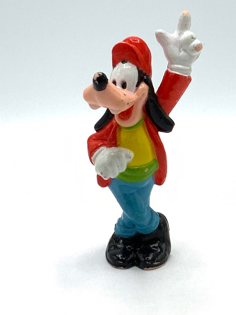 May include: A red, yellow, and blue plastic figurine of Goofy wearing a red baseball cap and a yellow shirt. He is standing with his right arm raised and his left hand on his hip.