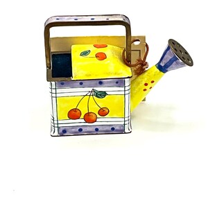 May include: A yellow metal watering can with a blue spout and handle. The can is decorated with a checkered pattern, red cherries, and yellow polka dots.
