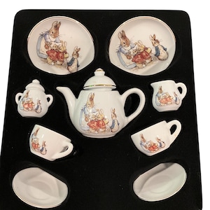 May include: A miniature porcelain tea set featuring Peter Rabbit illustrations. The set includes a teapot with a lid, two cups, two saucers, a creamer, and a sugar bowl with a lid, all decorated with colorful rabbit characters and gold trim.