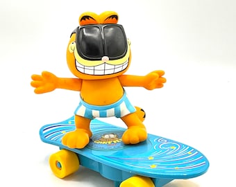 Puede incluir: Una figura de plástico de Garfield con gafas de sol y bañador azul y blanco a rayas está montada en una patineta azul con ruedas amarillas. La patineta tiene un diseño de olas azul y blanco con estrellas. El texto "Garfield" está impreso en la patineta.