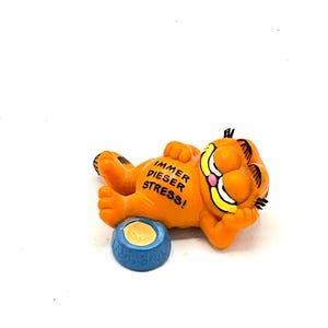 May include: Orange Garfield figurine lying on its back with its paws in the air. The figurine has a yellow bowl with a blue rim and the text "IMMER DIESER STRESS!" on its belly.