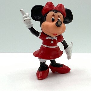 May include: A red and black plastic figurine of Minnie Mouse wearing a red dress and red shoes. She is holding her right arm up in the air with her index finger pointing up.