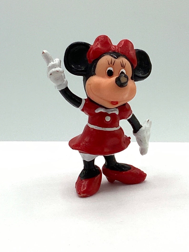 May include: A red and black plastic figurine of Minnie Mouse wearing a red dress and red shoes. She is holding her right arm up in the air with her index finger pointing up.