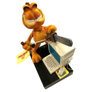 May include: Orange Garfield figurine holding a chainsaw, poised above a vintage computer. The figurine is on a black base, with a yellow tag featuring Garfield's name. The computer includes a keyboard, mouse, and monitor.